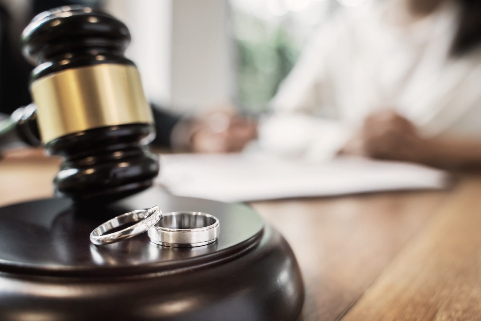 Close-up of a judge's gavel sitting on its block, with two wedding rings placed beside it. In the blurred background, two people are seated at a table, one holding a pen, suggesting a legal or courtroom setting, possibly related to divorce proceedings.