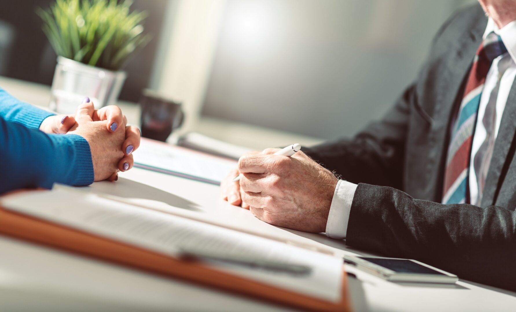 Two people are seated at a desk, engaging in a business conversation. One person's hands are clasped on the table, while the other holds a pen, ready to take notes on a document. A plant, some papers, and a phone are visible on the desk.