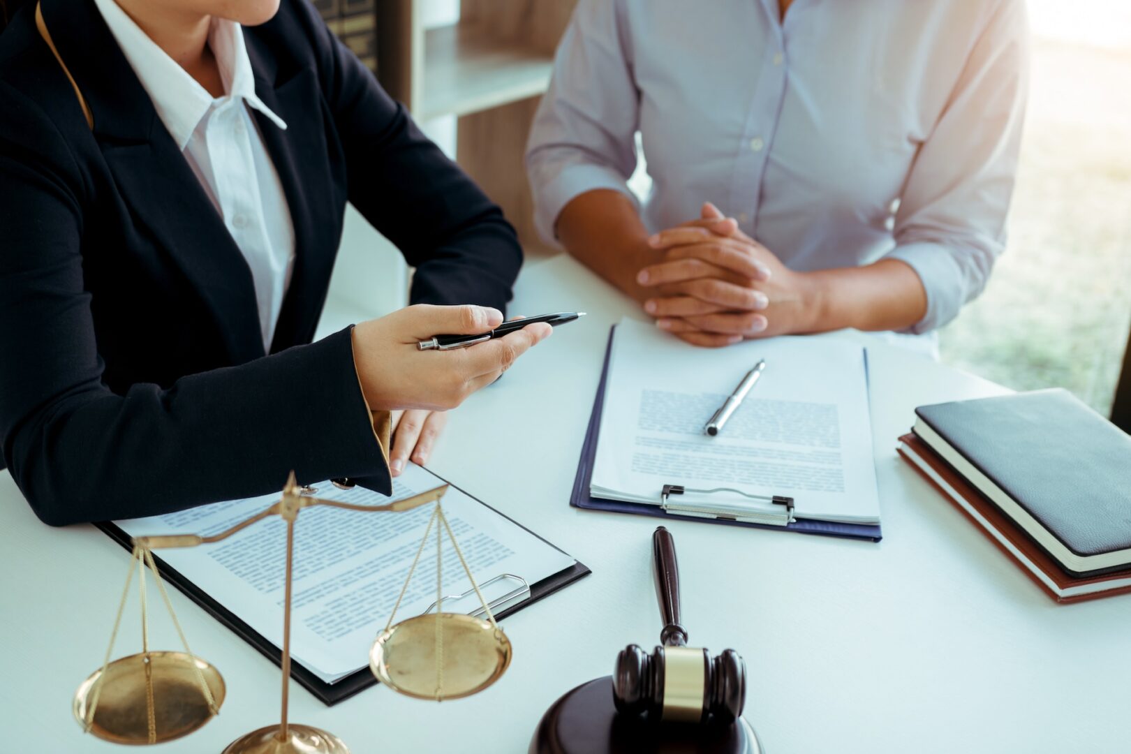 Two people are seated at a table with legal documents, a gavel, and scales of justice. One person is gesturing with a pen. The setting suggests a legal or business discussion.