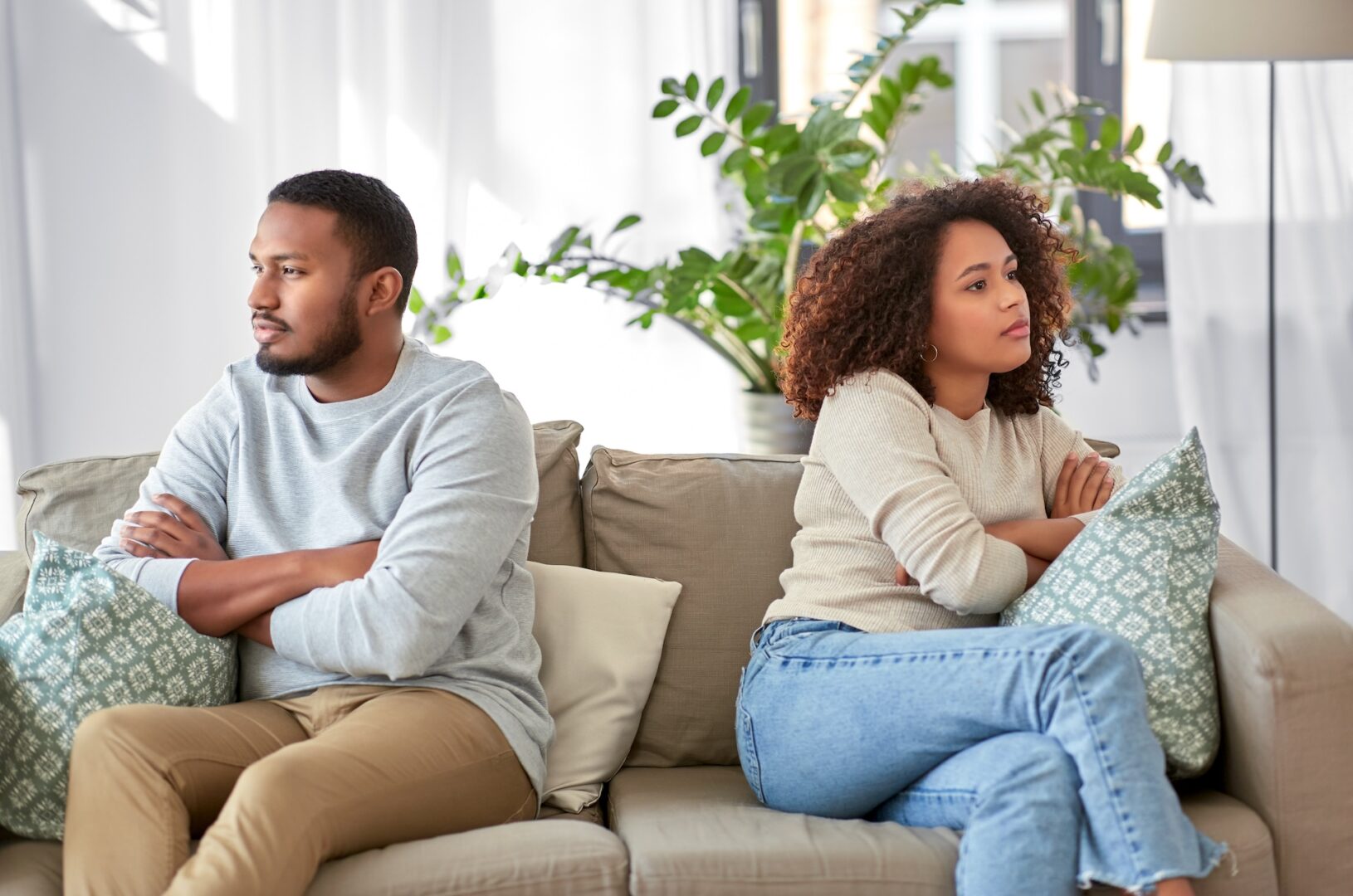 A man and a woman sit on a couch with their arms crossed, facing away from each other. They both look thoughtful and slightly displeased. There's a green plant in the background and decorative pillows on the couch.