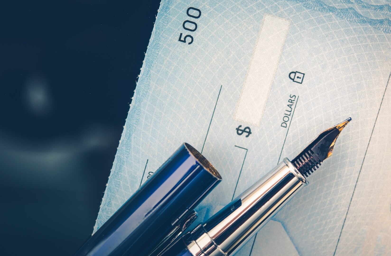 A close-up of a fountain pen resting on a blank check. The pen's cap is off, revealing the nib. The check is blue, showing various security features and a pre-printed number "500" near the top. The text "DOLLARS" is visible on the amount line.