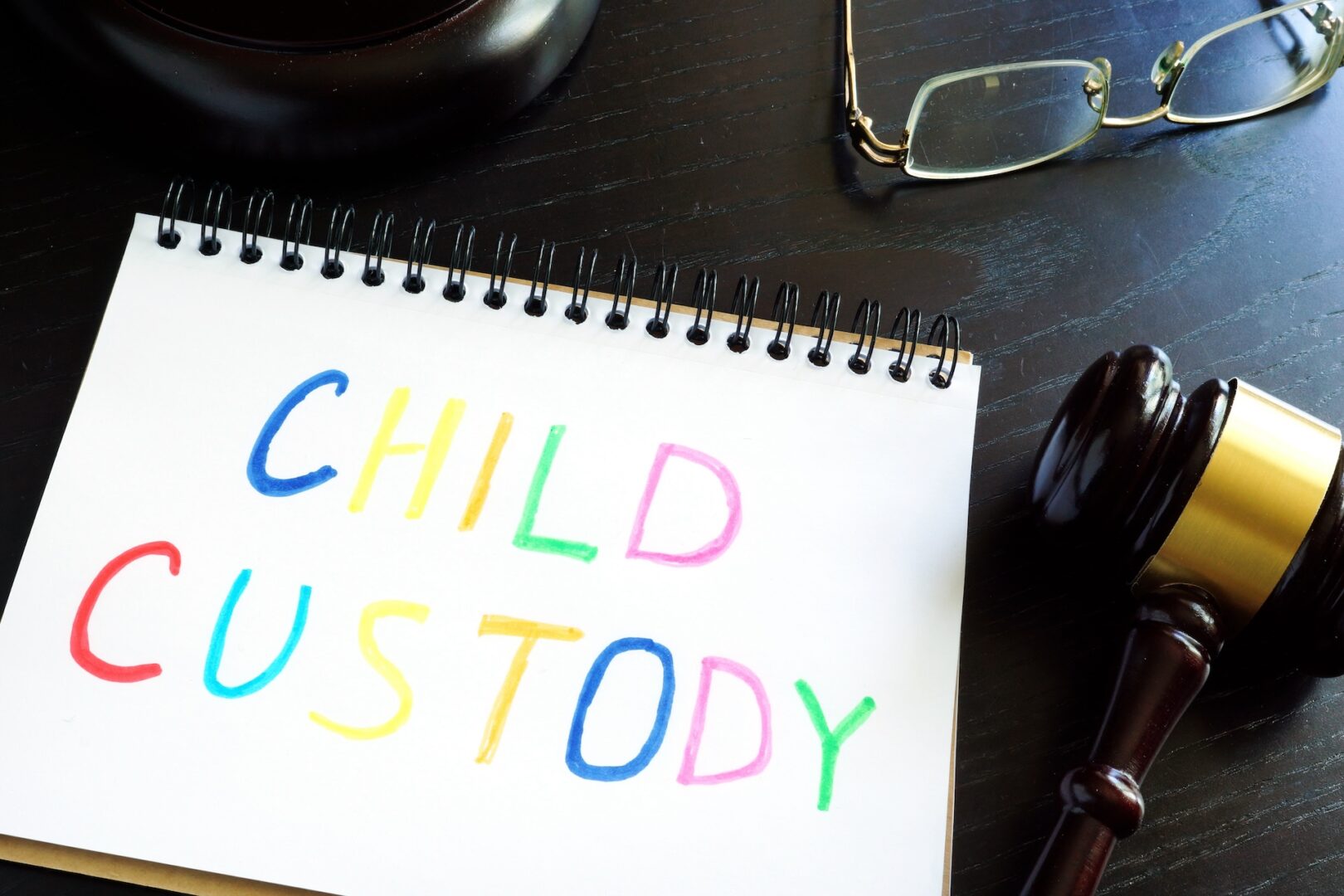 A notepad with "Child Custody" written in colorful letters lies on a dark wooden table. Nearby are a pair of eyeglasses and a judge's gavel, indicating a legal context.