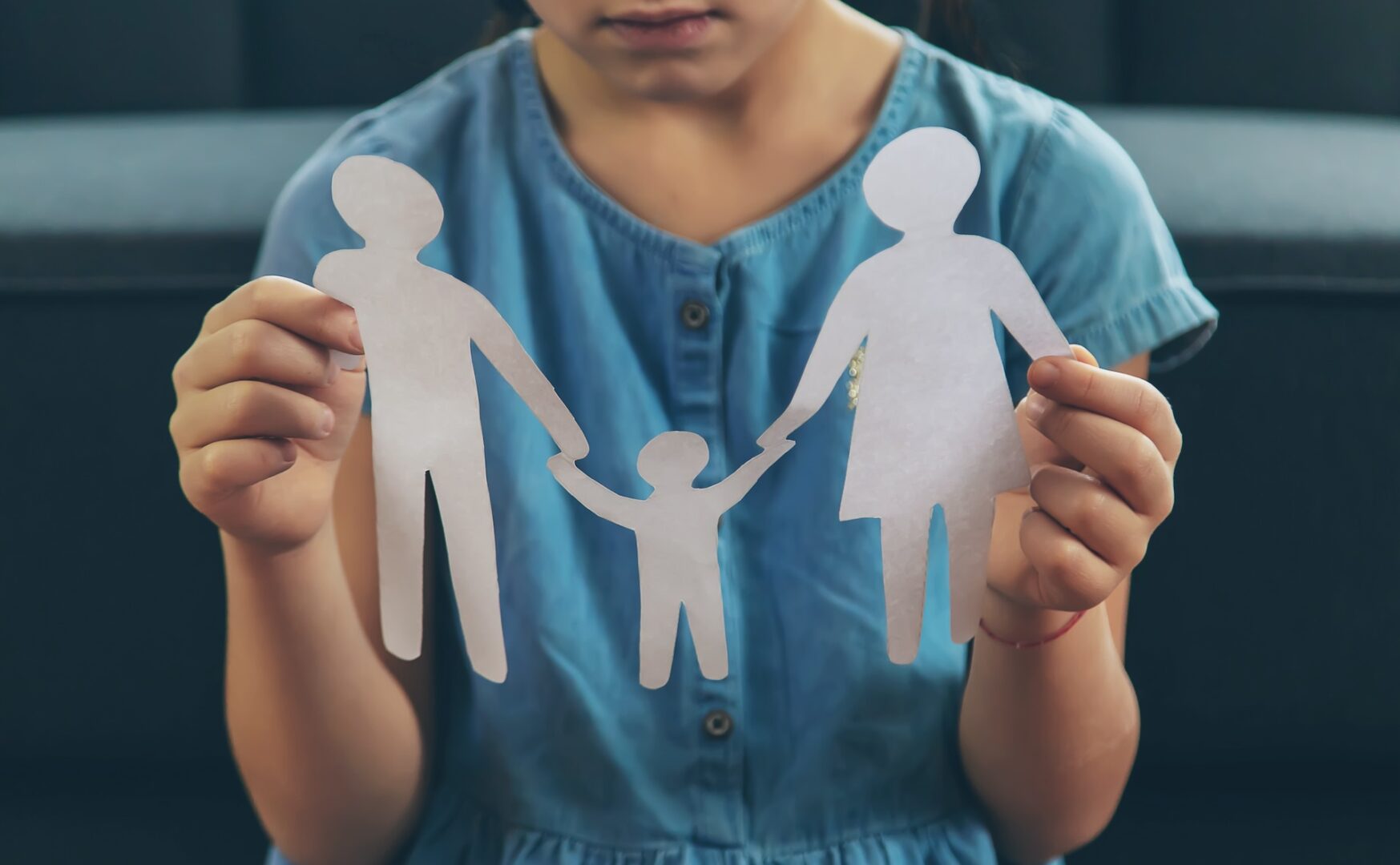 A child in a blue dress holds paper cutouts of a family—two adults and a child—symbolizing family unity or separation. The focus is on the cutouts, with the child partially visible in the background.