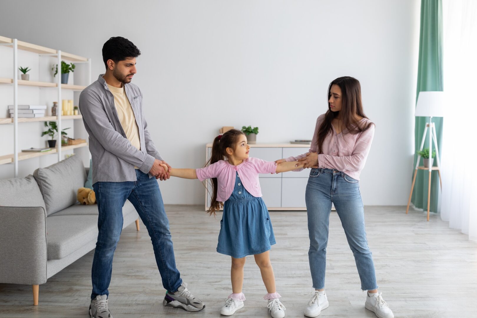 A young girl in a blue dress stands between a man and a woman, holding their hands. The man and woman face each other with neutral expressions. The room has a modern design with a sofa, shelves, and large window.