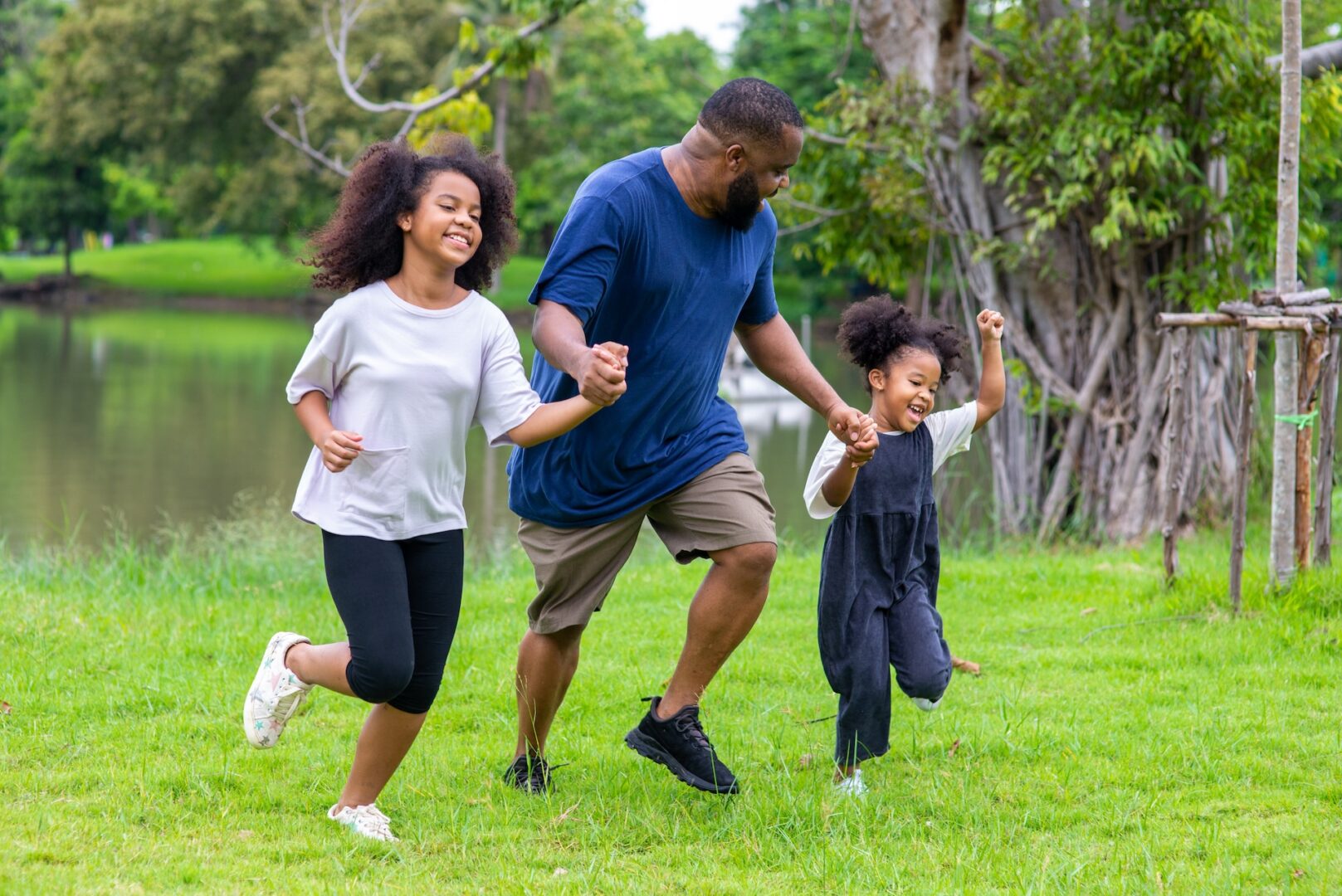 A father and his two daughters joyfully hold hands and run together on a grassy field near a pond. The older girl wears a white shirt and black pants, while the younger girl is in a white shirt with black overalls. The father wears a blue shirt.