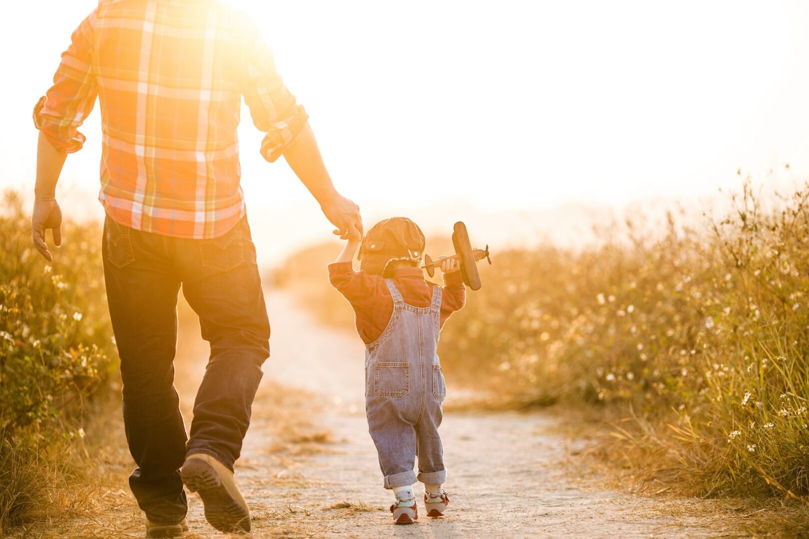 A parent and child walk down a sunlit path, hand in hand. The child, wearing overalls and a pilot hat, holds a toy airplane. Tall grass surrounds the path, and the sun sets in the background, casting a warm glow.