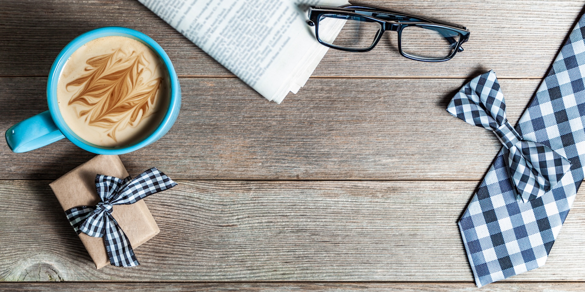 A wooden table displays a blue mug with a creamy coffee, a newspaper, black glasses, a small gift wrapped with a checkered ribbon, and a matching checkered necktie and bowtie. The scene conveys a stylish and cozy atmosphere.