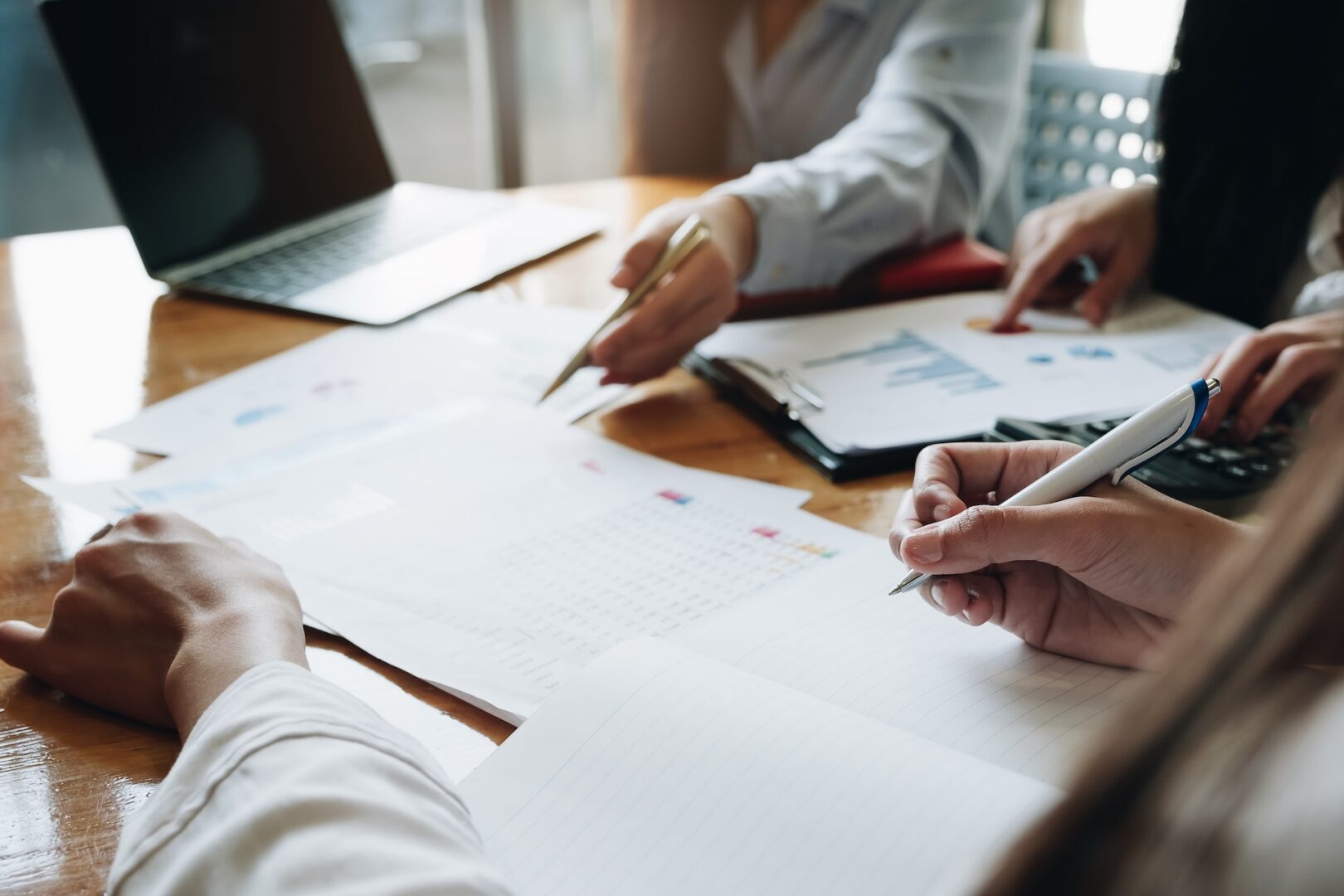 Three people work at a wooden table, reviewing documents and graphs. One person writes in a notebook, another points to a chart, and a laptop is open nearby. The scene conveys a collaborative business meeting or study session.