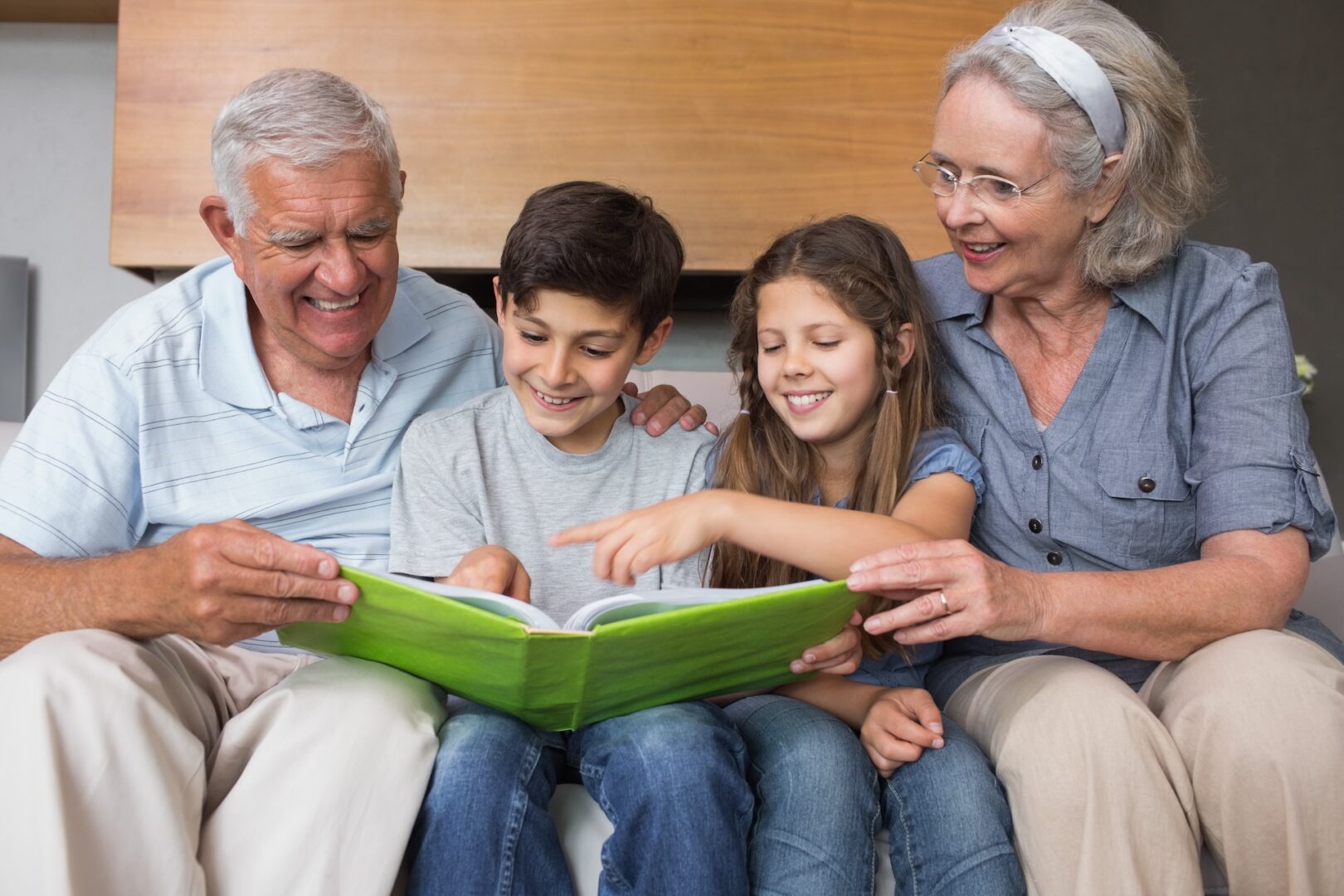 An elderly couple sits on a couch with two young children, a boy and a girl. They are all smiling and looking at a book that the grandfather is holding. The children appear engaged, and the grandmother is watching them fondly.