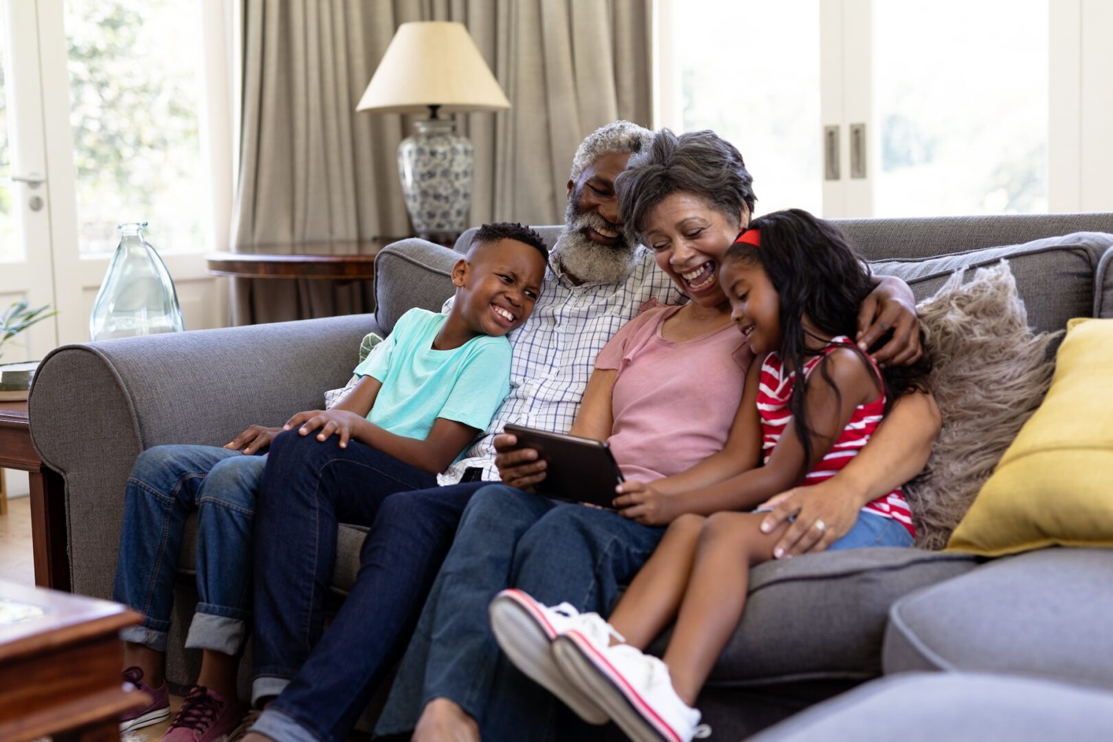 A joyful family sits together on a couch in a cozy living room. An elderly couple is in the center, with a young boy and girl on either side. They are all smiling and looking at a tablet, enjoying their time together.