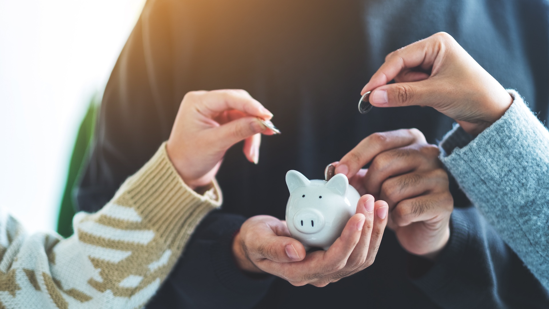 Three hands dropping coins into a white piggy bank held by a person wearing a black sweater. The scene suggests saving money collaboratively.