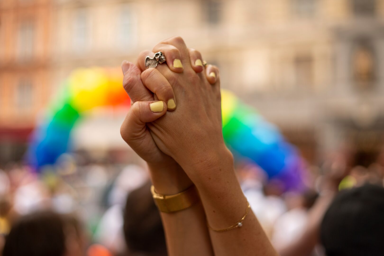 Two hands clasped together in a gesture of unity, with rainbow-themed decorations in the blurred background, symbolizing LGBTQ+ pride and solidarity.