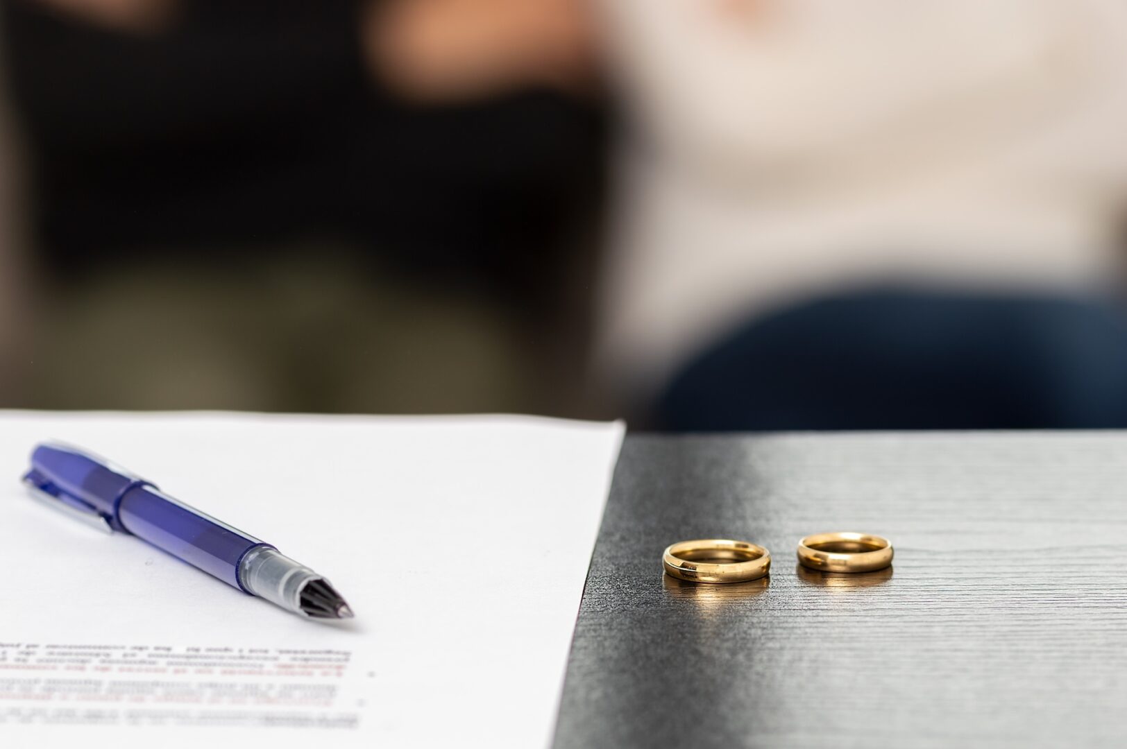 A pen rests on a legal document next to two gold wedding rings on a dark table. In the blurred background, two people sit with arms crossed, likely suggesting a separation or divorce context.