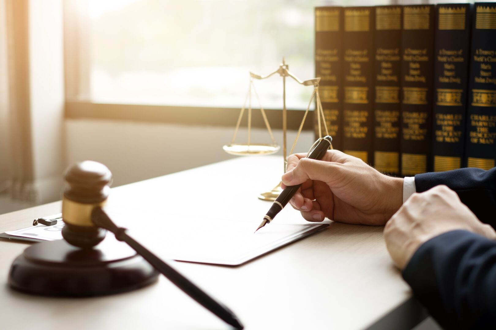 A person writes on a clipboard at a desk with a gavel to the left and a balanced scale in the background. Rows of law books line the back wall, and sunlight filters in through a window.