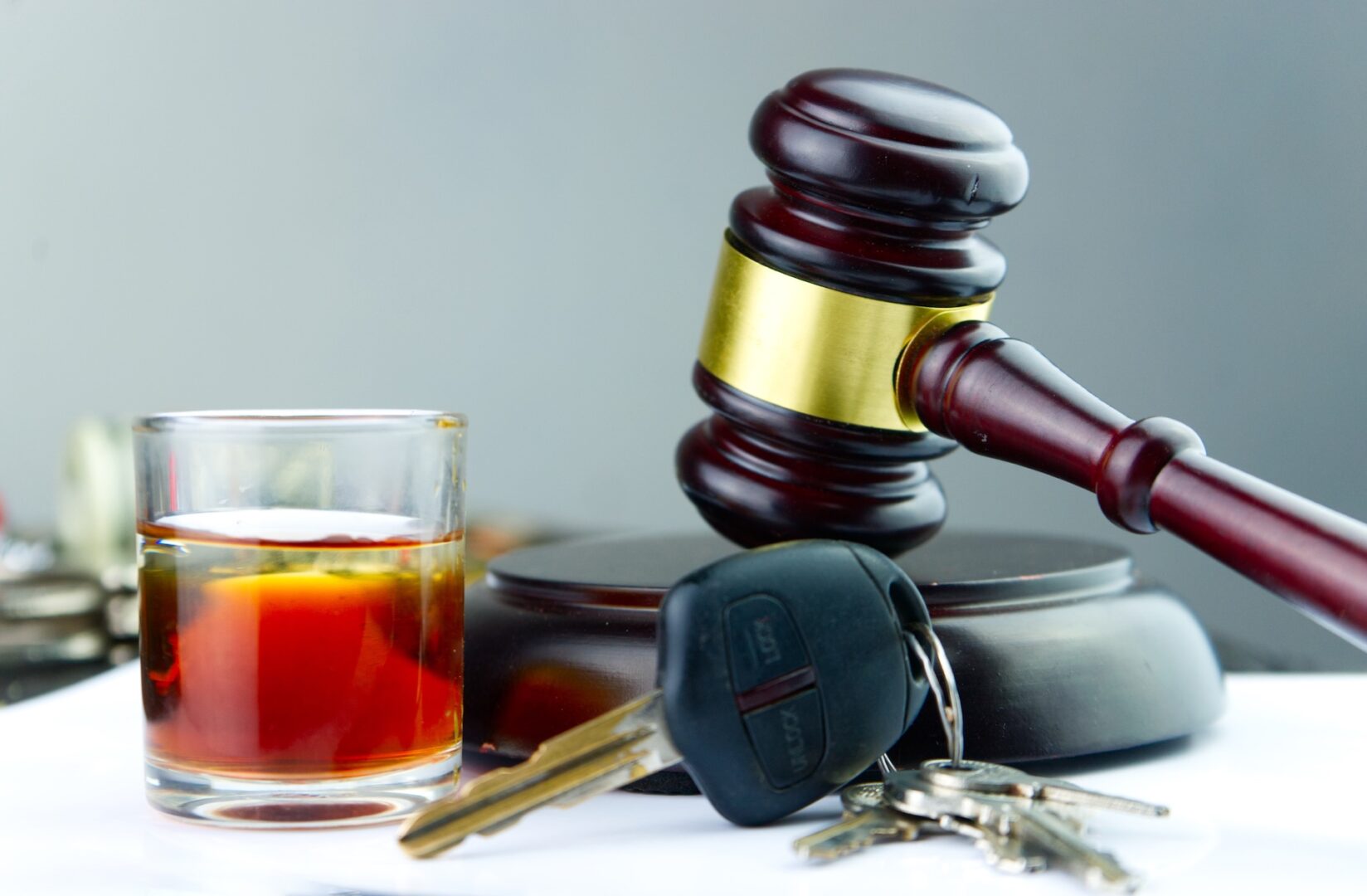 A gavel rests on its base next to a glass of amber liquid and a set of car keys on a surface, symbolizing legal consequences of drinking and driving. The background is blurred.