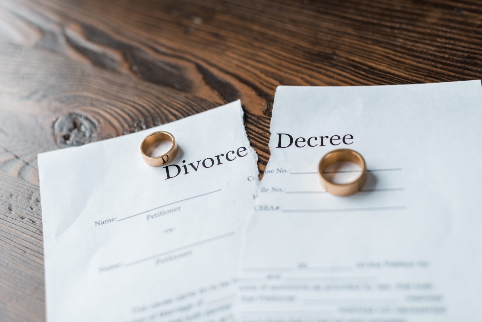 Two pieces of paper on a wooden table, torn apart, labeled "Divorce Decree." Each piece has a gold wedding ring placed on it, symbolizing the end of a marriage.