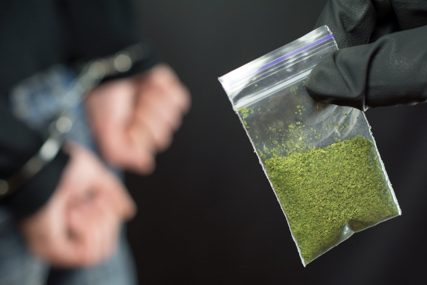 A gloved hand holds a small plastic bag containing a green powder, while a person in the background is handcuffed, suggesting a drug-related arrest.