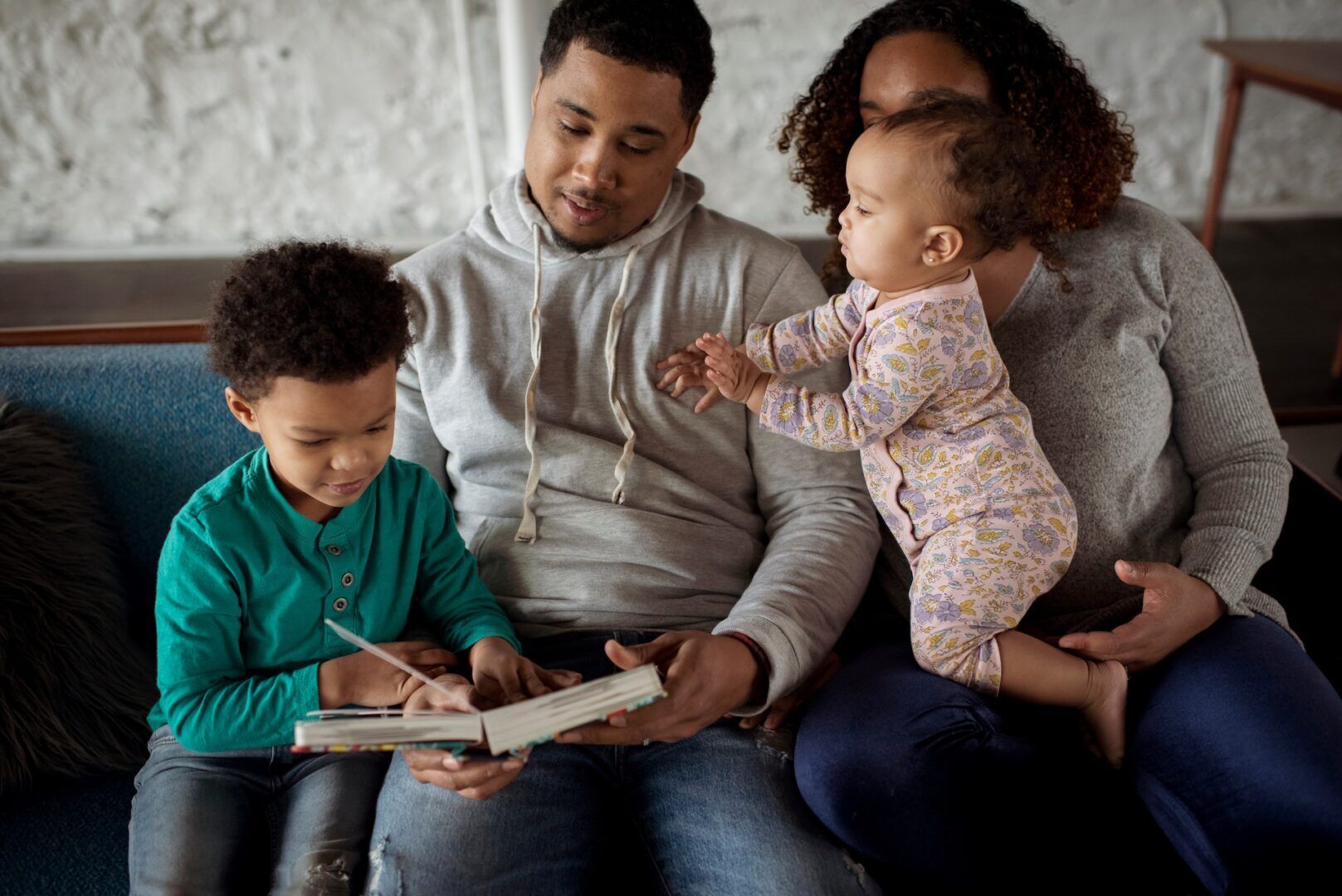 A man in a hoodie sits on a couch with a young child on his lap, reading a book together. A woman sits beside them holding a baby dressed in a patterned onesie, who is reaching toward the book. The family is engaged in a cozy indoor setting.