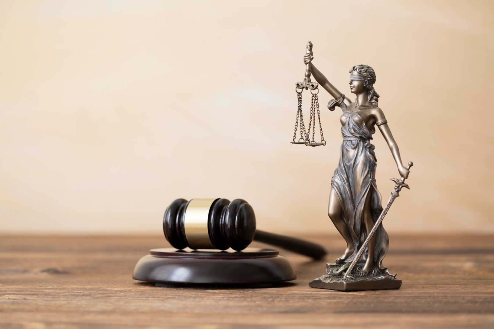 A bronze Lady Justice statuette holding scales and a sword stands beside a wooden gavel with a brass band, resting on its base. The background is a plain light brown surface, symbolizing law and justice.