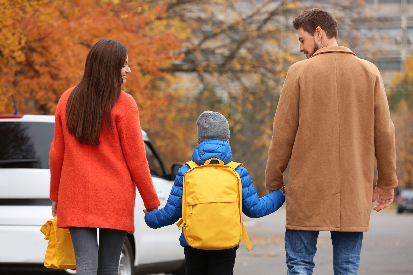 A woman in a red coat, a child in a blue jacket with a yellow backpack, and a man in a brown coat walk hand in hand on a street. Autumn leaves are on the trees, and a white car is parked nearby.