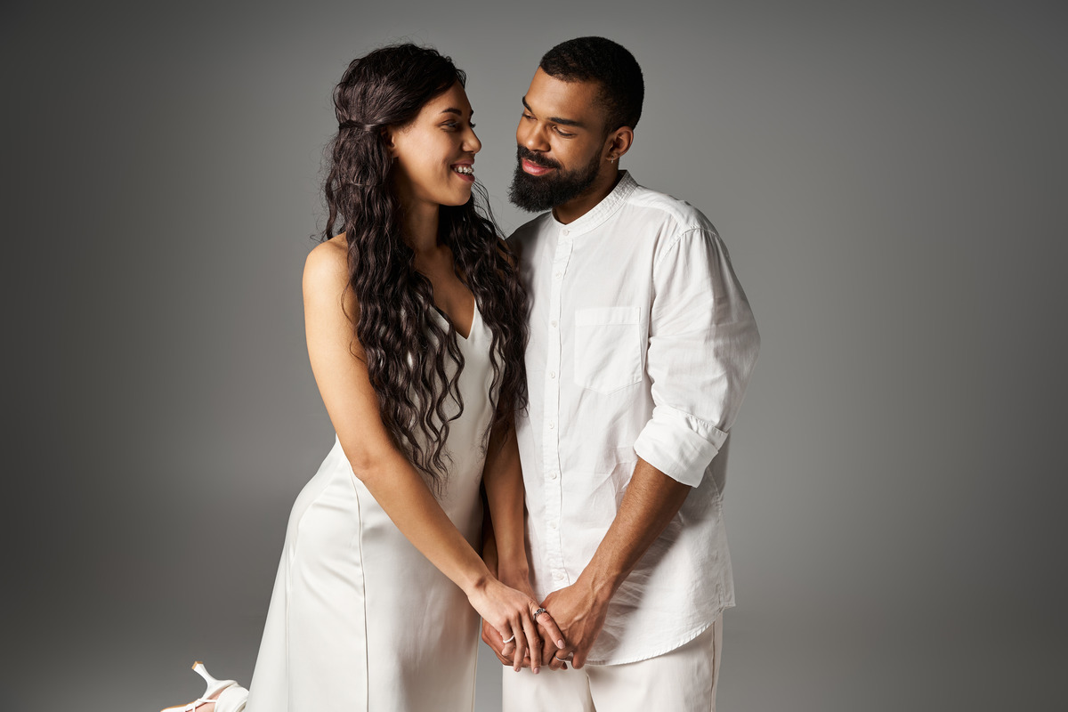 fashionable-couple-shares-a-tender-moment-in-style A young girl in a blue dress stands between a man and a woman, holding their hands. The man and woman face each other with neutral expressions. The room has a modern design with a sofa, shelves, and large window.